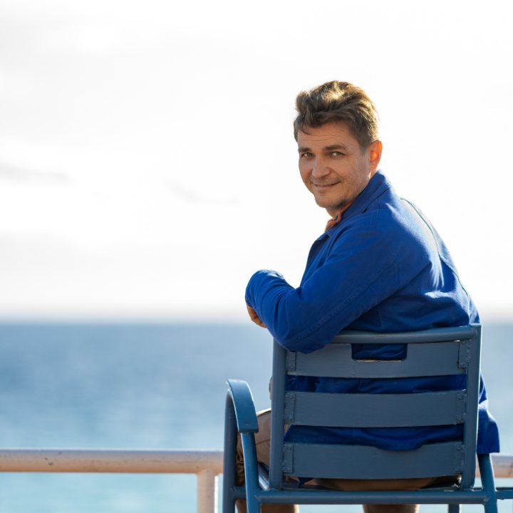 Alexei seated on a blue chair by the sea in Nice, looking back toward the camera.
